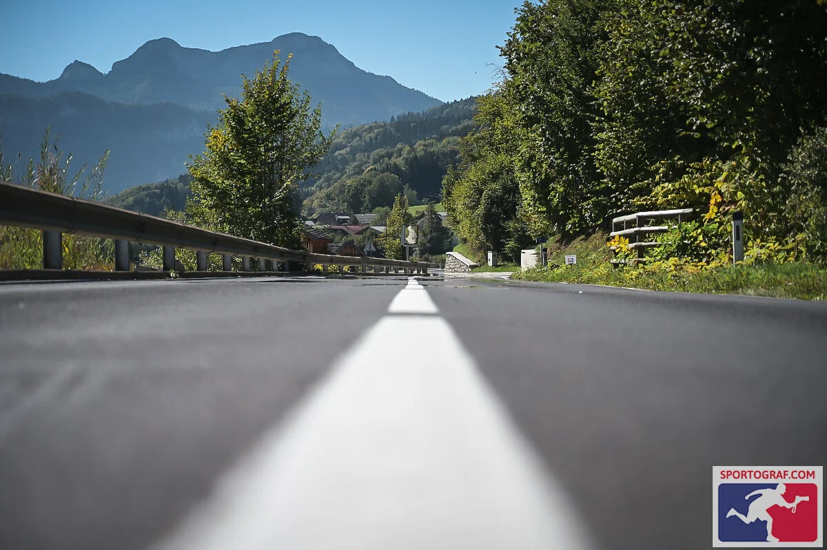 Blick aus Bodenhöhe auf die Straße am Attersee mit weißer Mittellinie, Leitplanke und Bergkulisse im Hintergrund